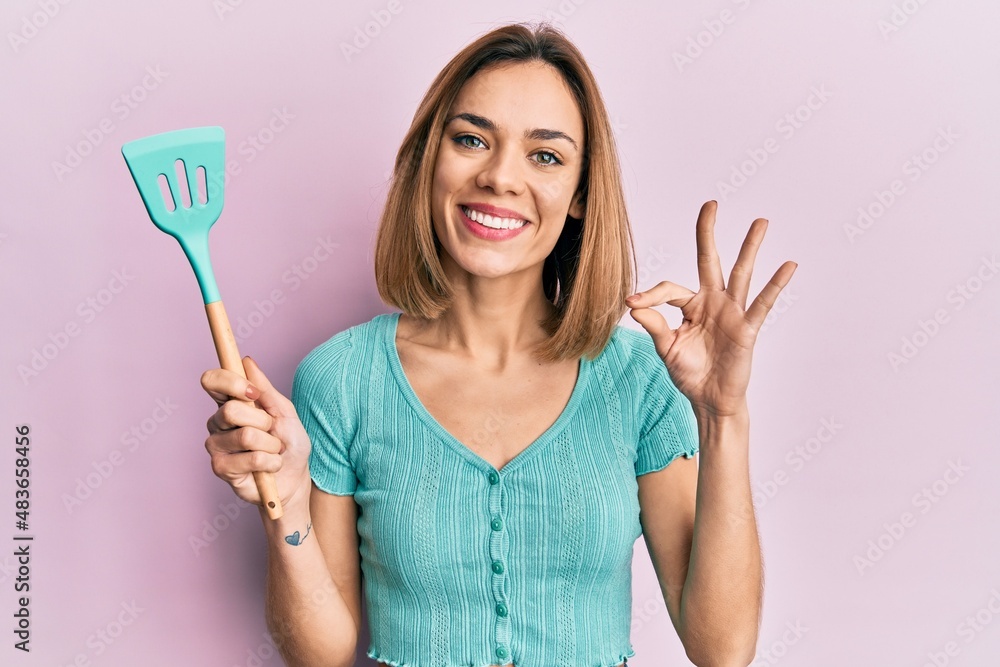 Young caucasian blonde woman holding silicone spatula doing ok sign with fingers, smiling friendly gesturing excellent symbol Young caucasian blonde woman holding silicone spatula doing ok sign with fingers, smiling friendly gesturing excellent symbol