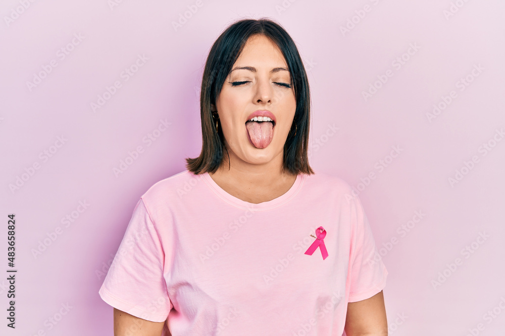 Young hispanic woman wearing pink cancer ribbon on t shirt sticking tongue out happy with funny expression. emotion concept. Young hispanic woman wearing pink cancer ribbon on t shirt sticking tongue out happy with funny expression. emotion concept.