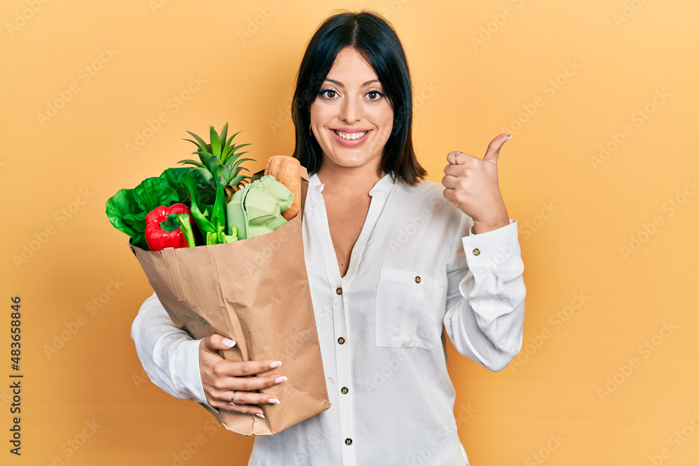 Young hispanic woman holding paper bag with groceries smiling happy and positive, thumb up doing excellent and approval sign Young hispanic woman holding paper bag with groceries smiling happy and positive, thumb up doing excellent and approval sign
