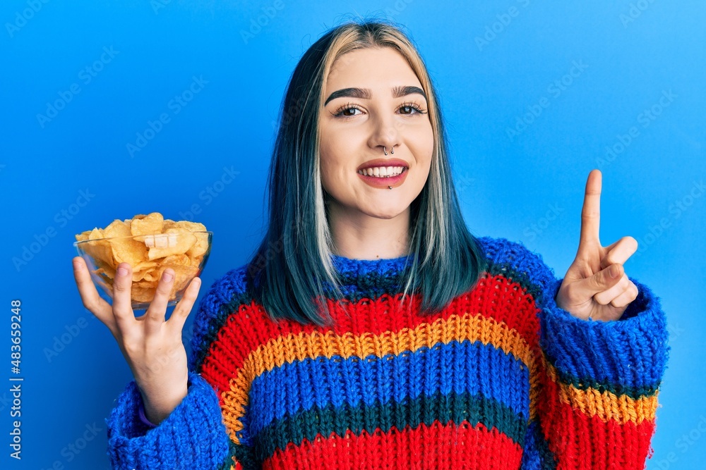 Young modern girl holding potato chips smiling with an idea or question pointing finger with happy face, number one Young modern girl holding potato chips smiling with an idea or question pointing finger with happy face, number one