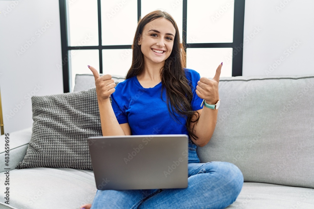 Young brunette woman using laptop at home success sign doing positive gesture with hand, thumbs up smiling and happy. cheerful expression and winner gesture. Young brunette woman using laptop at home success sign doing positive gesture with hand, thumbs up smiling and happy. cheerful expression and winner gesture.