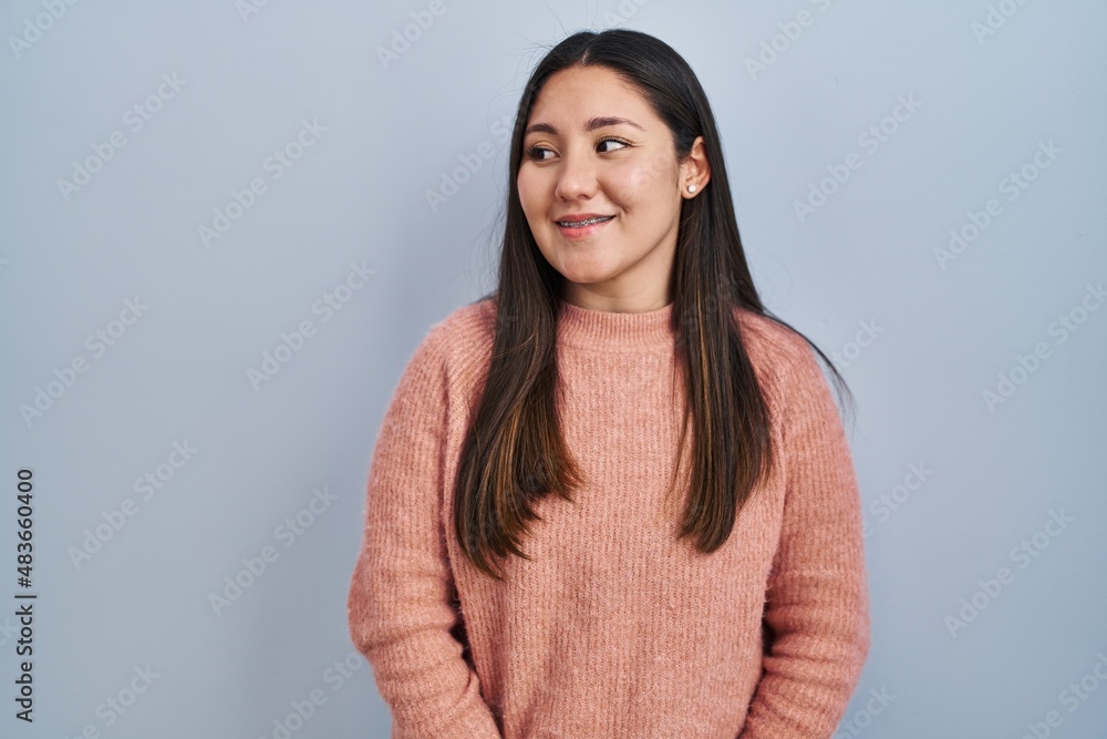 Young latin woman standing over blue background looking away to side with smile on face, natural expression. laughing confident. Young latin woman standing over blue background looking away to side with smile on face, natural expression. laughing confident.