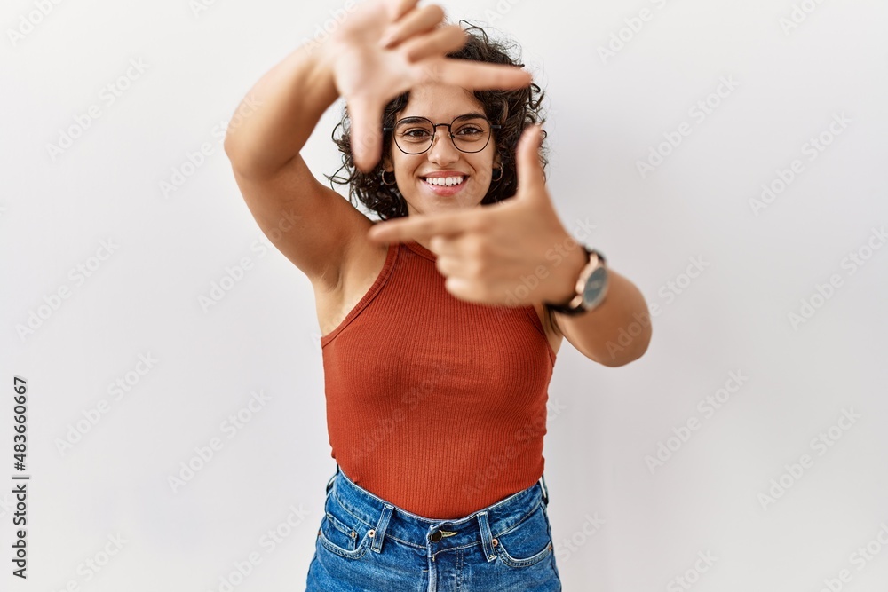 Young hispanic woman wearing glasses standing over isolated background smiling making frame with hands and fingers with happy face. creativity and photography concept. Young hispanic woman wearing glasses standing over isolated background smiling making frame with hands and fingers with happy face. creativity and photography concept.