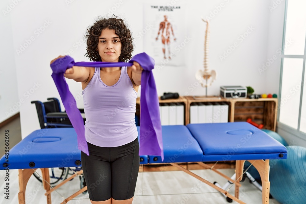 Fototapeta premium Young hispanic woman doing rehabilitation using elastic band at clinic