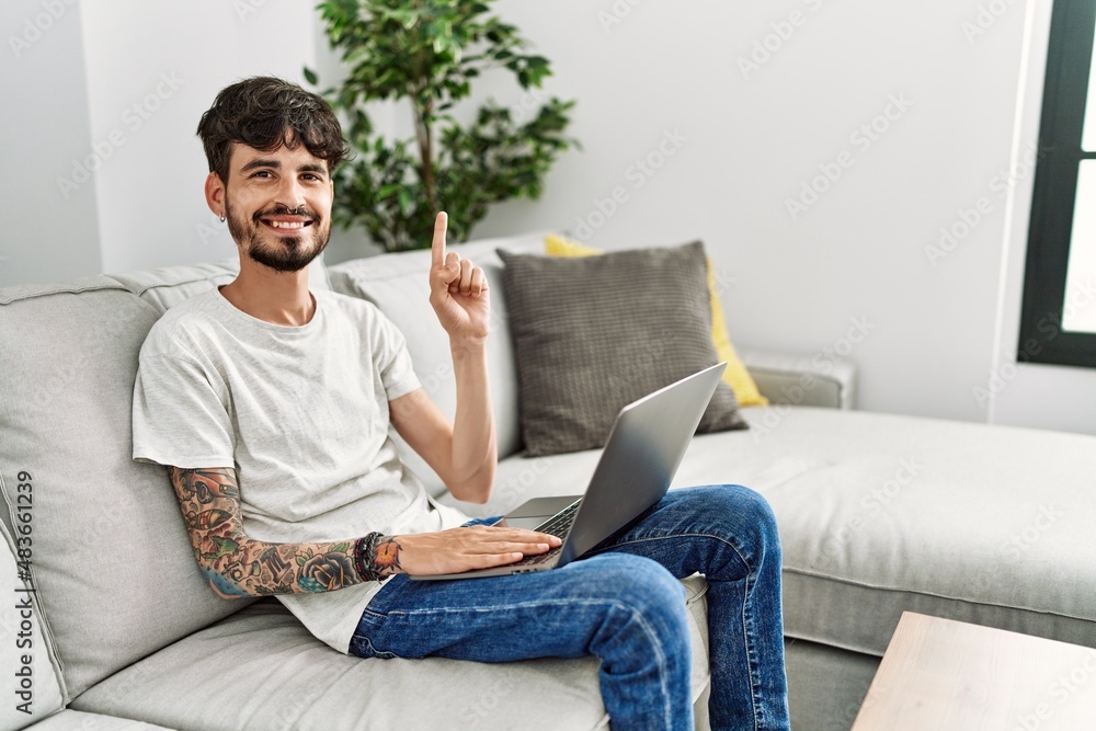 Hispanic man with beard sitting on the sofa showing and pointing up with finger number one while smiling confident and happy. Hispanic man with beard sitting on the sofa showing and pointing up with finger number one while smiling confident and happy.