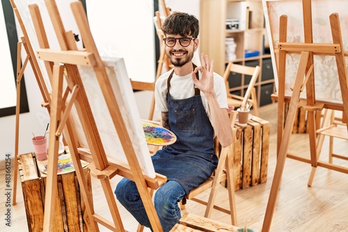 Hispanic man with beard at art studio doing ok sign with fingers, smiling friendly gesturing excellent symbol