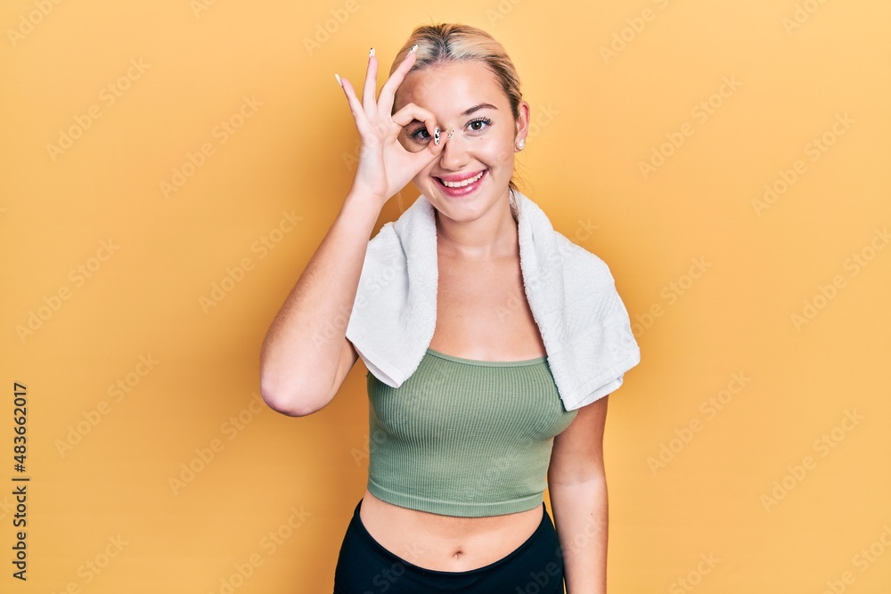 Young blonde girl wearing sportswear and towel doing ok gesture with hand smiling, eye looking through fingers with happy face. Young blonde girl wearing sportswear and towel doing ok gesture with hand smiling, eye looking through fingers with happy face.