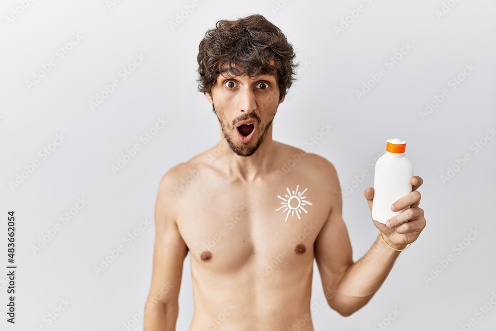 Fototapeta premium Young hispanic man standing shirtless holding sunscreen lotion in shock face, looking skeptical and sarcastic, surprised with open mouth