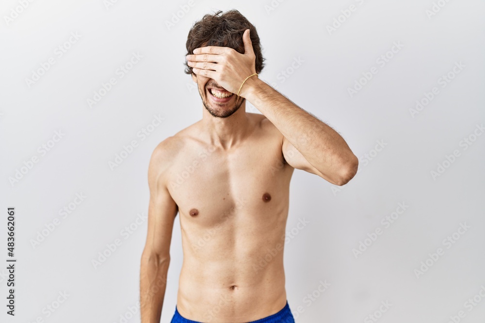 Fototapeta premium Young hispanic man standing shirtless over isolated, background smiling and laughing with hand on face covering eyes for surprise. blind concept.