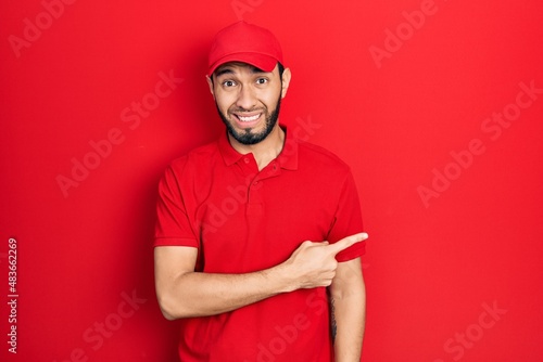 Hispanic man with beard wearing delivery uniform and cap pointing aside worried and nervous with forefinger  concerned and surprised expression