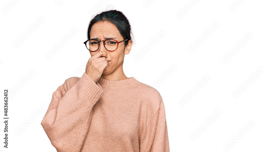 Young hispanic girl wearing casual clothes and glasses smelling something stinky and disgusting, intolerable smell, holding breath with fingers on nose. bad smell