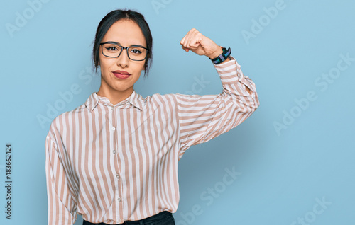 Young hispanic girl wearing casual clothes and glasses strong person showing arm muscle, confident and proud of power