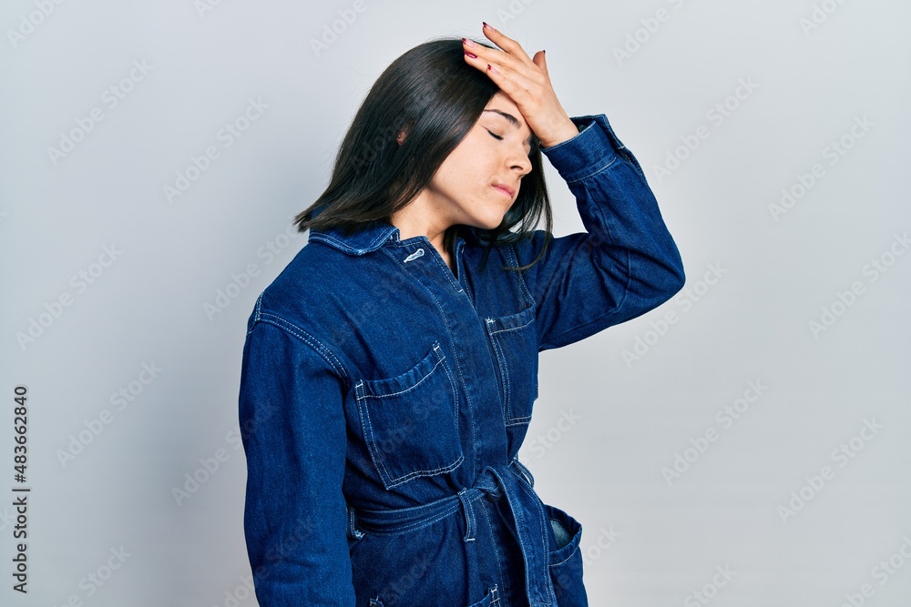 Young brunette woman wearing denim jumpsuit surprised with hand on head for mistake, remember error. forgot, bad memory concept. Young brunette woman wearing denim jumpsuit surprised with hand on head for mistake, remember error. forgot, bad memory concept.
