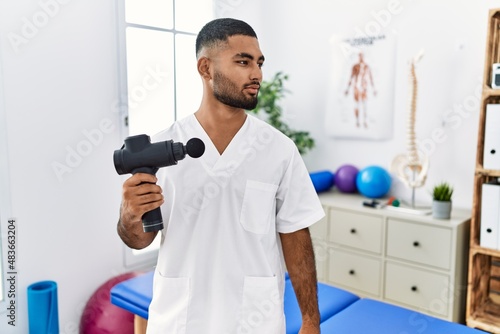 Young indian physiotherapist holding therapy massage gun at wellness center looking to side, relax profile pose with natural face and confident smile.