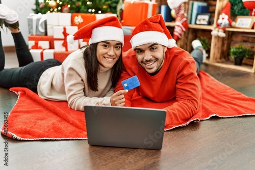 Young hispanic couple using laptop and credit card lying on the floor by christmas tree at home.