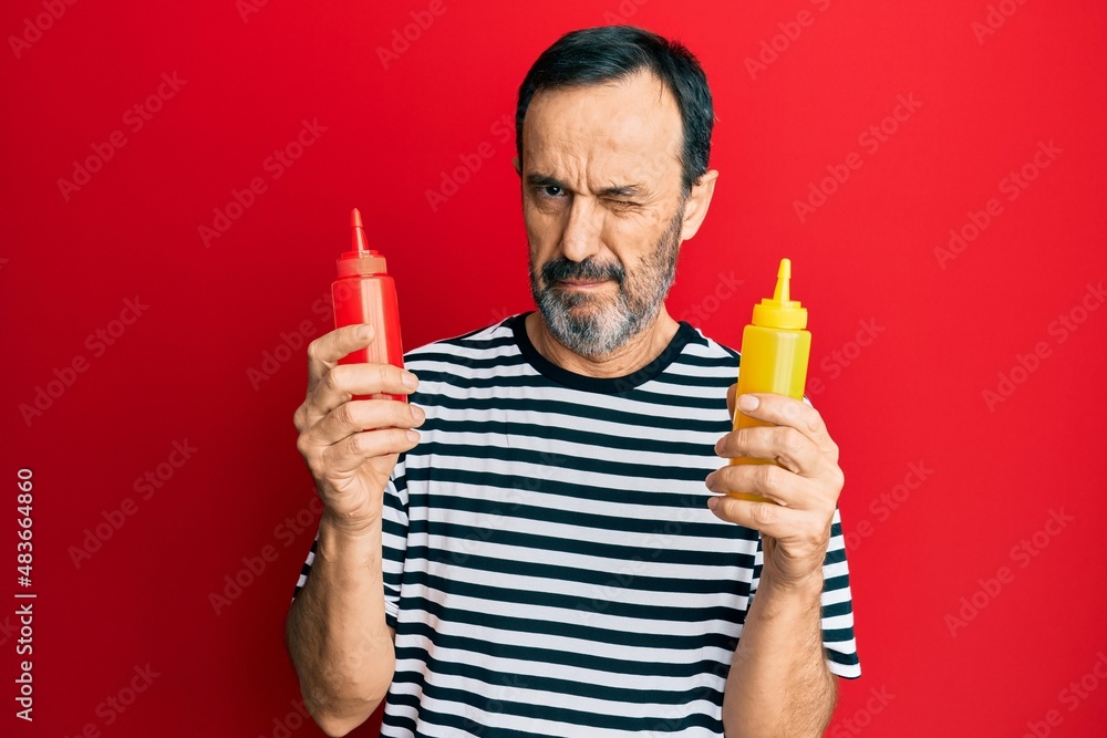 Middle age hispanic man holding ketchup and mustard bottle skeptic and nervous, frowning upset because of problem. negative person. Middle age hispanic man holding ketchup and mustard bottle skeptic and nervous, frowning upset because of problem. negative person.