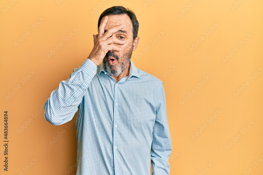 Middle age hispanic man wearing casual clothes peeking in shock covering face and eyes with hand, looking through fingers afraid