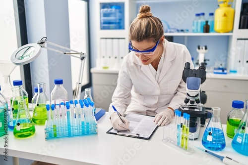 Young blonde woman wearing scientist uniform writing on clipboard at laboratory