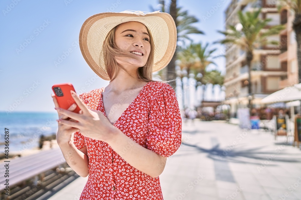 Fototapeta premium Young chinese girl wearing summer hat using smartphone at the promenade.