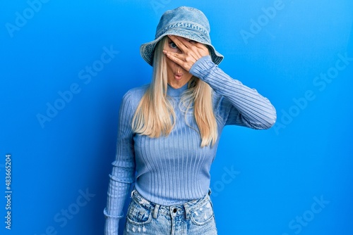 Young blonde woman wearing casual denim hat peeking in shock covering face and eyes with hand, looking through fingers with embarrassed expression.