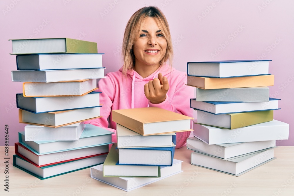 Young caucasian woman sitting on the table with books beckoning come here gesture with hand inviting welcoming happy and smiling Young caucasian woman sitting on the table with books beckoning come here gesture with hand inviting welcoming happy and smiling