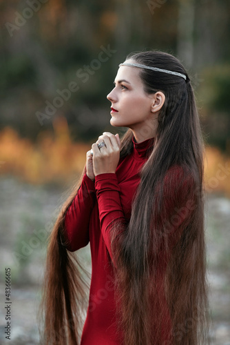 Mystical portrait of a long-haired sorceress praying in an autumn forest. Beautiful young woman in a red dress, posing prettily against a sunset forest backdrop