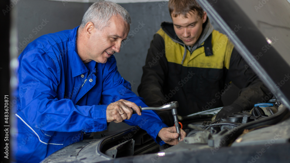 Senior experienced mechanic teaches his colleague how to repair a car Senior experienced mechanic teaches his colleague how to repair a car