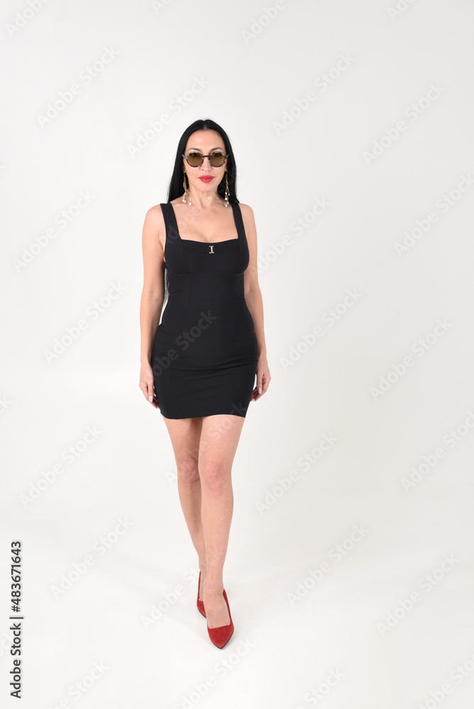 A brunette girl is beautiful and stylish in a black dress and dark glasses with bare feet in the studio on a white isolated background A brunette girl is beautiful and stylish in a black dress and dark glasses with bare feet in the studio on a white isolated background