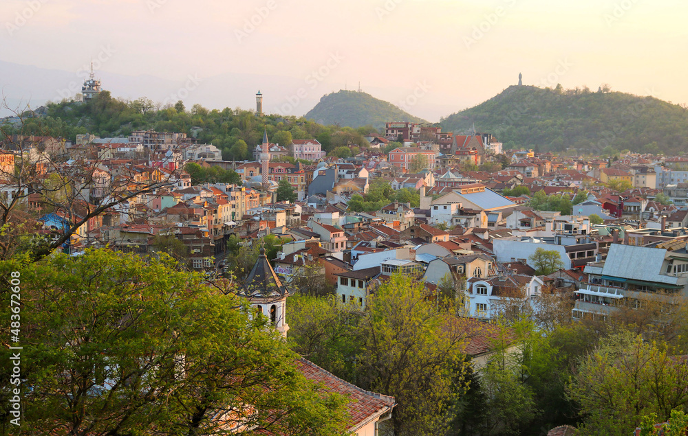 Tourist postcard of the ancient Plovdiv in Bulgaria with a panoramic view of the city with buildings and monuments, trees, hills and mountains in the background, during the sunset