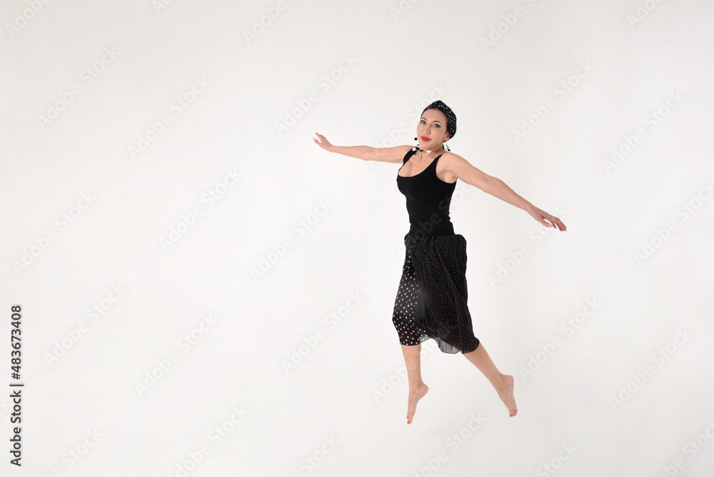 A brunette woman in a black dress barefoot jumped into the air in delight on a white background