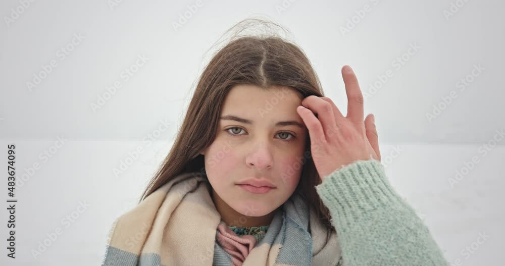 Teenager adjusting hair in winter. Young female in warm clothes touching long dark hair and looking at camera against snowy field and gray sky in winter countryside Teenager adjusting hair in winter. Young female in warm clothes touching long dark hair and looking at camera against snowy field and gray sky in winter countryside