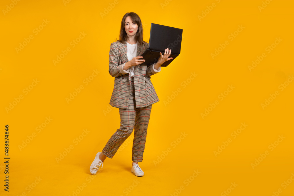 Woman with a laptop looks into camera. Businesswoman on a yellow background. Girl with a laptop computer. Businesswoman uses a computer. Concept - laptops for business. Fashionable businesswoman Woman with a laptop looks into camera. Businesswoman on a yellow background. Girl with a laptop computer. Businesswoman uses a computer. Concept - laptops for business. Fashionable businesswoman
