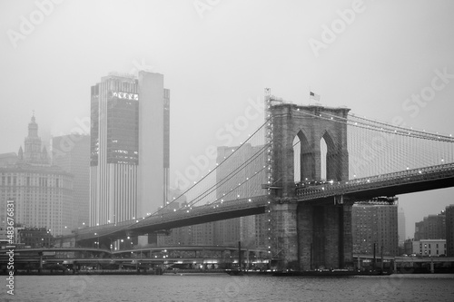 Black and white photo of skyscrapers of Manhattan and Brooklyn bridge on foggy and cloudy day. Famous bridge. Postcard view of New York, USA. © Maria Sbytova
