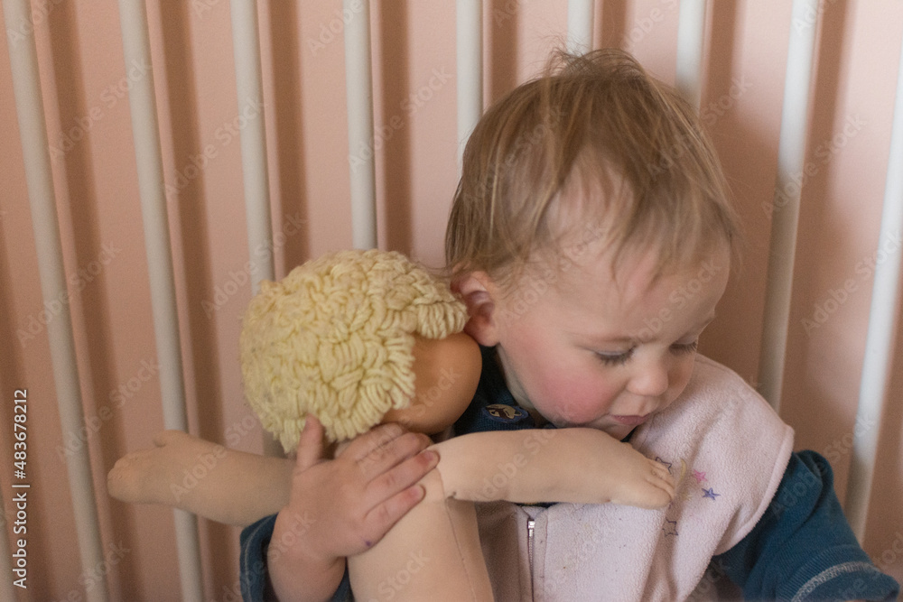Toddler wakes from nap and hugs her baby doll; child in wearable blanket for safe sleeping Toddler wakes from nap and hugs her baby doll; child in wearable blanket for safe sleeping