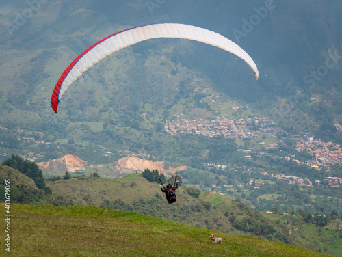 Men Flying in a Red Paraglider near the Mountains while a Small Dog Runs Through the Green Field in Belmira, Antioquia, Colombia photo