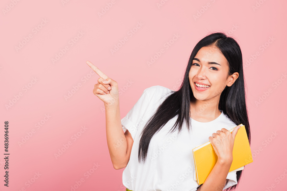 Portrait of happy Asian beautiful young woman confident smiling holding orange book open pointing finger to side copy space, studio shot isolated on pink background, education concept Portrait of happy Asian beautiful young woman confident smiling holding orange book open pointing finger to side copy space, studio shot isolated on pink background, education concept
