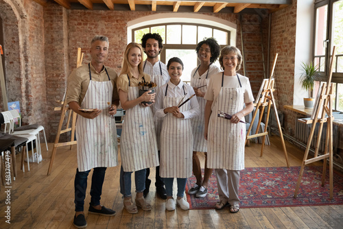 Canvas Multiethnic group of happy diverse art school students in craft artistic studio wearing aprons, holding paintbrushes, standing together, looking at camera