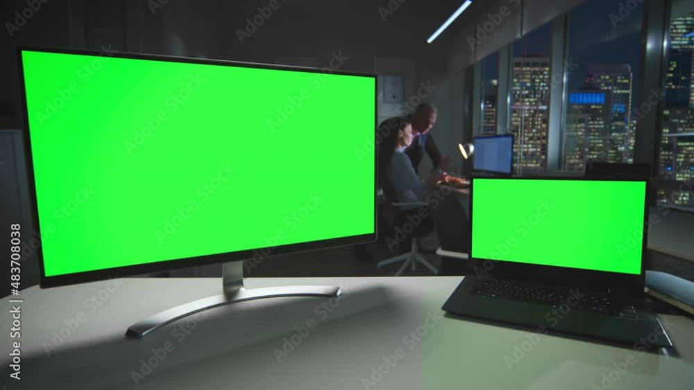 Computer screen and laptop with green screen in dark office with night city view through windows downtown Computer screen and laptop with green screen in dark office with night city view through windows downtown