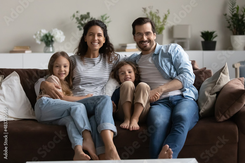 Happy homeowners well-being family portrait, bank loan and medical insurance cover concept. Beautiful couple and two preschool children sitting on cozy sofa in living room smiling looking at camera