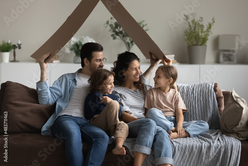 Young loving couple their preschool cute children sitting on cozy sofa in living room holding carton like roof  symbol of bank loan  new house owners  relocation day  happy homeowners family  concept