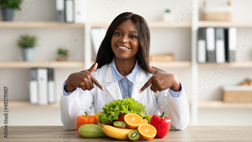 Portrait of happy african american dietitian pointing at fresh fruits and vegetables  smiling at camera at clinic