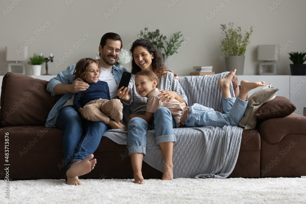 Happy family with kids have fun use new mobile application on cellphone. Couple and siblings sit on sofa in living room hold smartphone looks at screen make videocall, selfie, play videogame concept