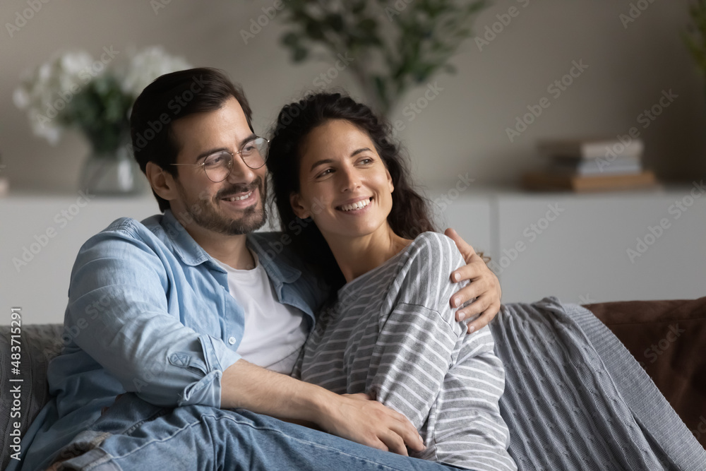 Dreamy beautiful couple in love embracing sit on sofa at home looking into distance daydreaming about bright future, enjoy romantic date feeling bond. Homeowners family, new house, relations concept Dreamy beautiful couple in love embracing sit on sofa at home looking into distance daydreaming about bright future, enjoy romantic date feeling bond. Homeowners family, new house, relations concept