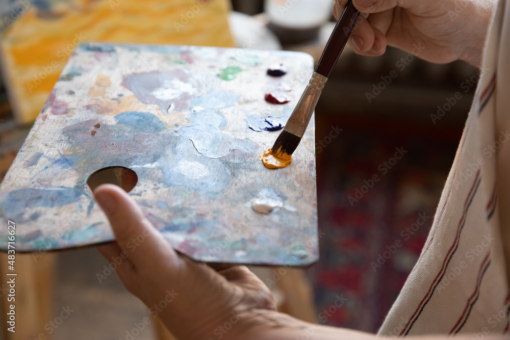 Artist holding rectangular palette, using paintbrush, blending paints, mixing yellow and blue colors, working in studio. Art school student hand with brush and pallet. Drawing tools concept Artist holding rectangular palette, using paintbrush, blending paints, mixing yellow and blue colors, working in studio. Art school student hand with brush and pallet. Drawing tools concept