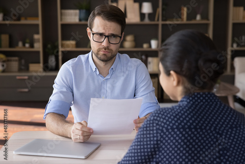 Unhappy young male hr manager in eyeglasses holding paper CV resume, feeling unsure about female Indian candidate. Unskilled woman job seeker making bad first impression on employer at office meeting