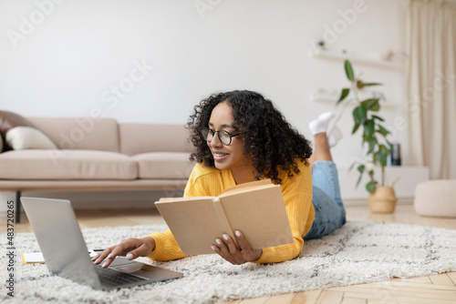 Young black woman reading book near laptop, getting ready for exam or working on project, lying on floor at home