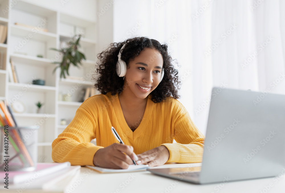 Happy African American woman in headphones having online meeting on laptop, writing down info during distance lesson Happy African American woman in headphones having online meeting on laptop, writing down info during distance lesson