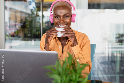 African woman drinking coffee while using laptop in coffee shop - Digital nomad and freelance lifestyle concept photo