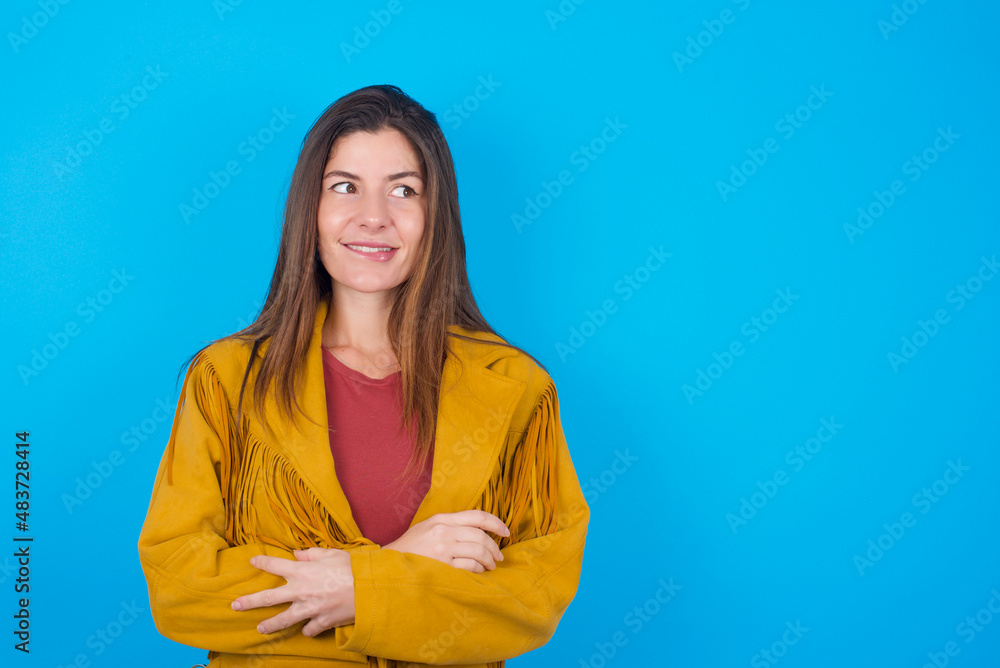 Fototapeta premium young brunette woman wearing yellow fringed jacket over blue background with thoughtful expression, looks away keeps hands down bitting his lip thinks about something pleasant.
