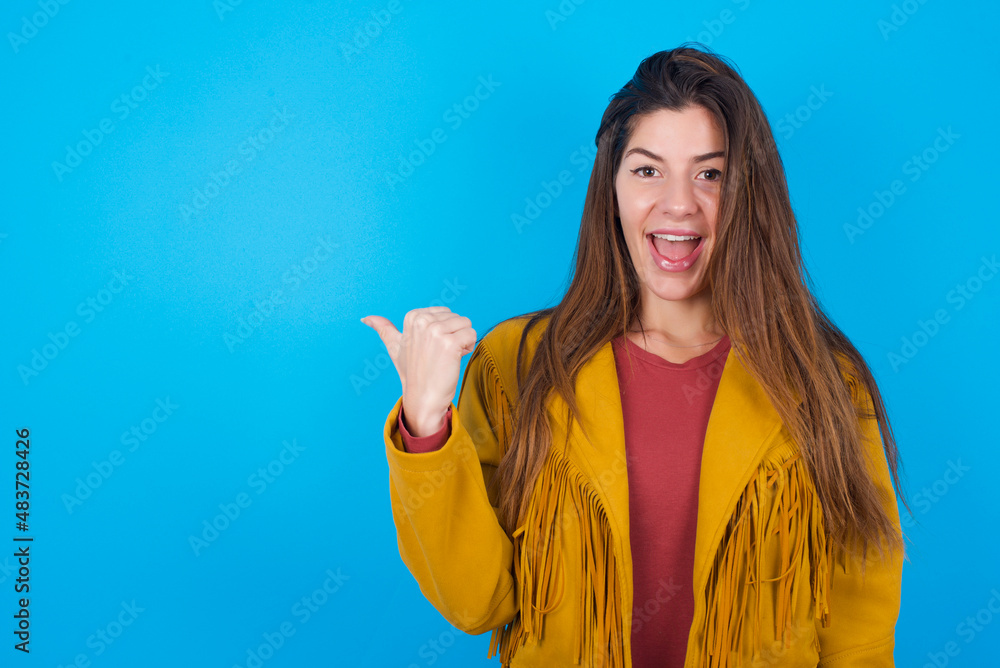 Stupefied young brunette woman wearing yellow fringed jacket over blue with surprised expression, opens eyes and mouth widely, points aside with thumb, shows something strange. Advertisement concept. Stupefied young brunette woman wearing yellow fringed jacket over blue with surprised expression, opens eyes and mouth widely, points aside with thumb, shows something strange. Advertisement concept.